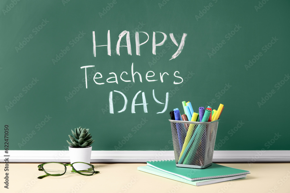 Text HAPPY TEACHERS DAY on green chalkboard with markers holder, eyeglasses and notebooks in classroom Text HAPPY TEACHERS DAY on green chalkboard with markers holder, eyeglasses and notebooks in classroom