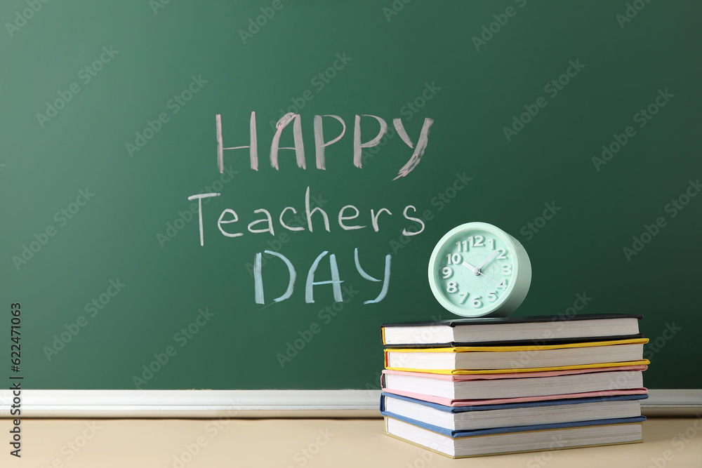 Text HAPPY TEACHERS DAY on green chalkboard with alarm clock and books in classroom Text HAPPY TEACHERS DAY on green chalkboard with alarm clock and books in classroom