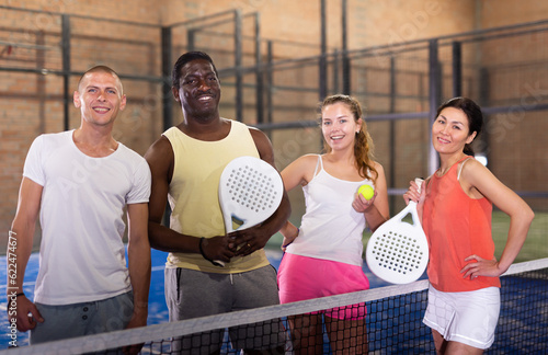 Happy smiling men and women of different nationalities in sportswear with rackets and balls in hands posing near net on indoor padel court. © JackF