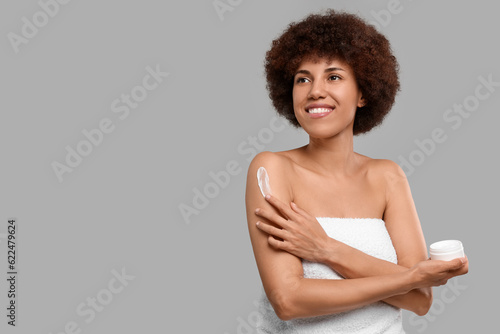 Beautiful young woman applying body cream onto arm on grey background, space for text