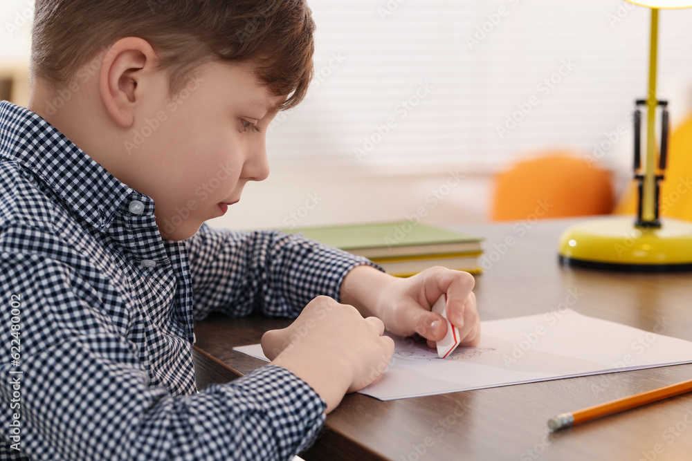 Little boy erasing mistake in his notebook at wooden desk indoors Little boy erasing mistake in his notebook at wooden desk indoors