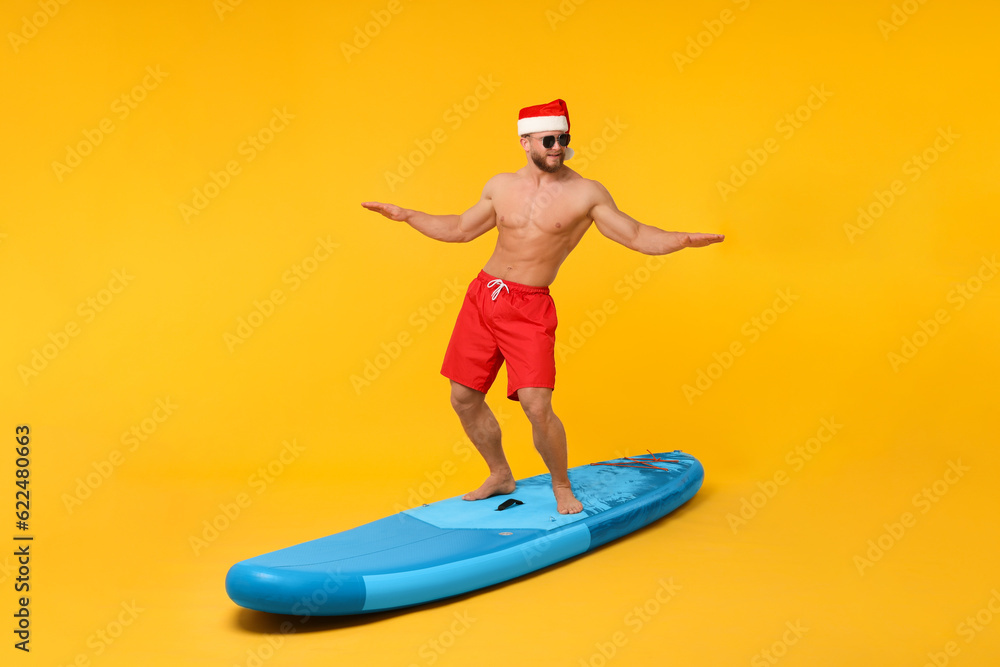 Happy man in Santa hat balancing on SUP board against orange background
