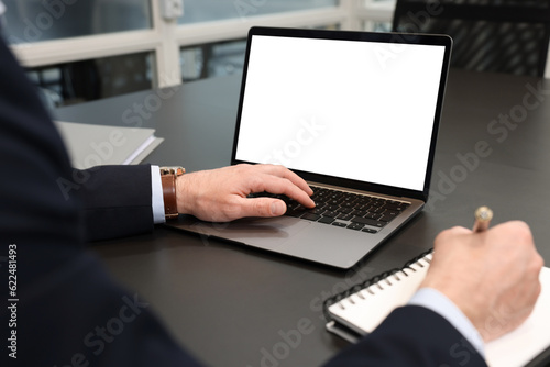 Man writing notes while working on laptop at black desk in office, closeup