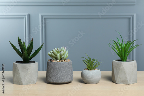 Many different artificial plants in flower pots on wooden table near grey wall
