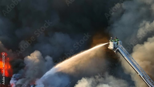 Cinematic drone shot of two firefighters on ladder spraying water on burning building while dark toxic fumes rising up - Aerial top down zoom shot