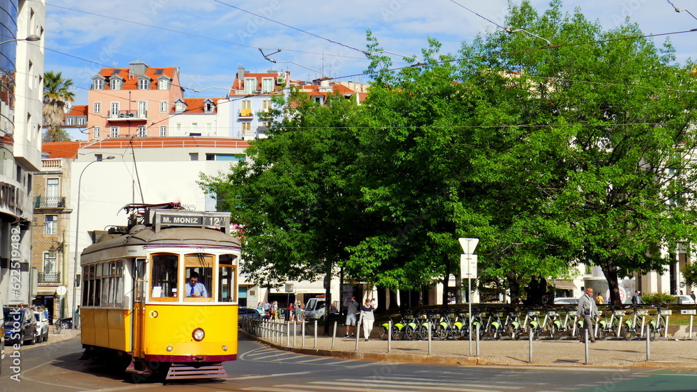 Naklejka premium gelbe nostalgische Straßenbahn in Lissabon fährt neben grünen Bäumen bei Sonne durchs Zentrum