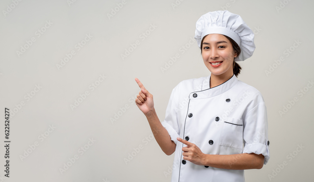 Smiling Young beautiful asian woman chef in uniform pointing finger to copy space isolated background. Cooking woman Occupation chef or baker People in kitchen restaurant and hotel. Smiling Young beautiful asian woman chef in uniform pointing finger to copy space isolated background. Cooking woman Occupation chef or baker People in kitchen restaurant and hotel.