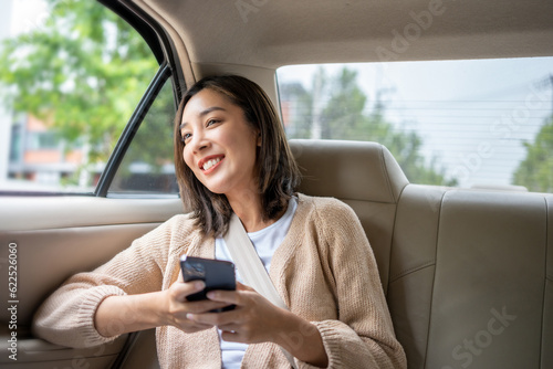 Relaxing moment of beautiful woman sitting in car back seats using smartphone play social media with safety belt and look out the window. Female happy in car traveling on the road to destination. © Chanakon