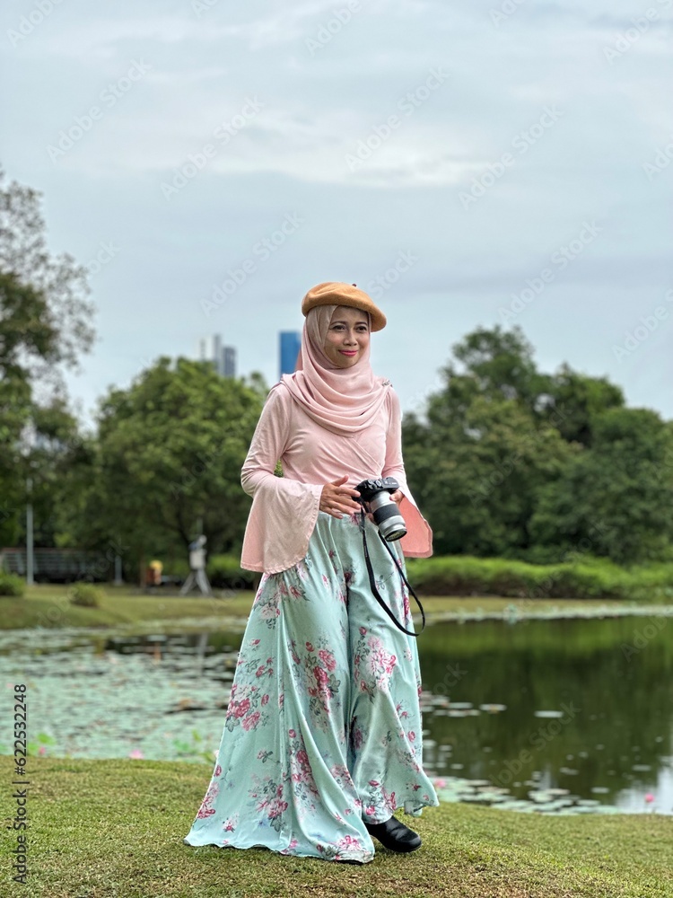 Portrait of beautiful young Muslim girl wearing Hijab and floral dress in spring outdoor scenes holding camera. Stylish Muslim female hijab fashion lifestyle portraiture concept.