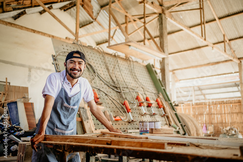 Asian entrepreneur in a hat smiling at the camera standing in a woodcraft workshop