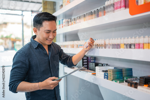 male shopkeeper looking at the digital tablet while checking the liquid stock at the shelf photo