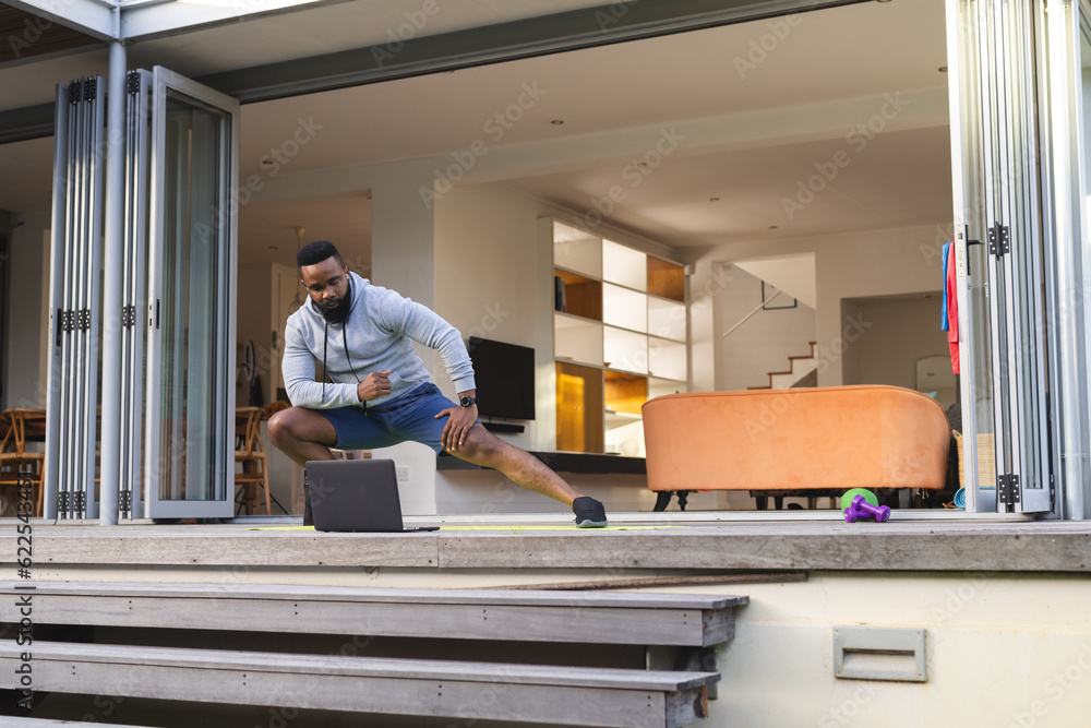 Focused african american man exercising and stretching on mat with laptop in garden Focused african american man exercising and stretching on mat with laptop in garden