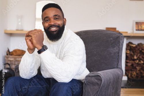 Portrait of happy african american man wearing watch, white jumper and sitting on armchair at home photo