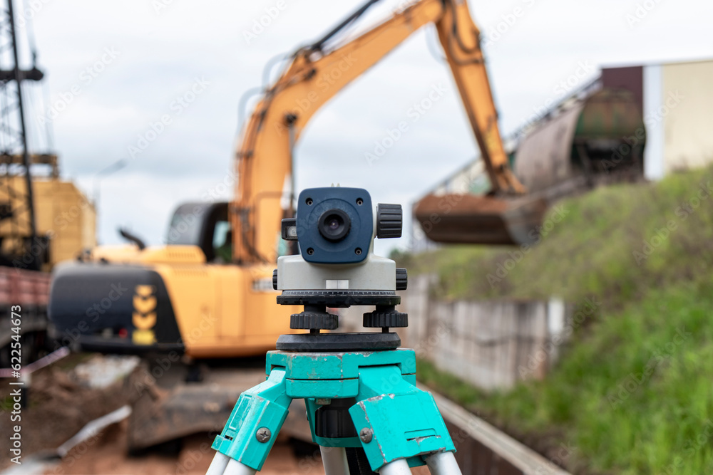 Level with an excavator in the background. Construction level or theodolite. Geodetic instruments and equipment for the construction and earthworks.