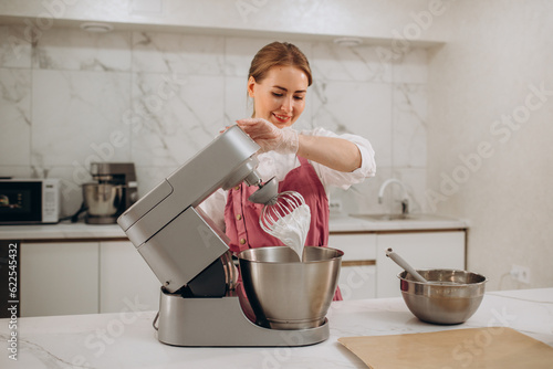 Close up of female chef holding egg beater with whipped cream. Metal bowls with sweet delicious cream on background.