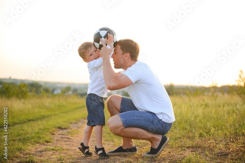 Fototapeta Naklejka Na Ścianę i Meble -  father with a little son plays football on the green grass in the park. Happy family having fun and playing football on a green grassy lawn on a sunny day. Family concept, Father's Day