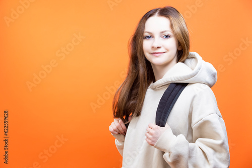 Winsome Caucasian Smiling Teenager Girl in Hoody Jacket Posing with Backpack Indoors Against Orange . photo