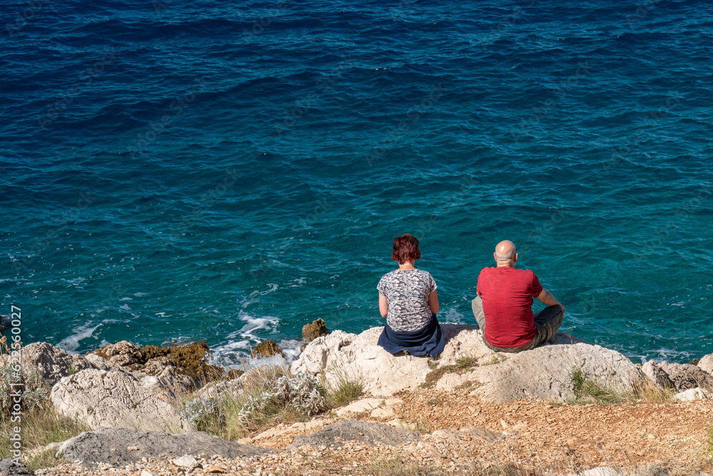 .A man and a woman sit on a rocky seashore and enjoy the beautiful view of the blue, transparent sea. Vacation and trave