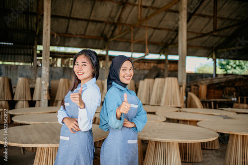 two female entrepreneurs stand with their backs to each other and show their thumbs up at a woodcraft shop