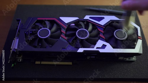 A computer technician takes apart a dirty video card to remove dust with compressed air and a brush. Cooling fans are spinning on the video card, the light illuminates the coolers from the inside.