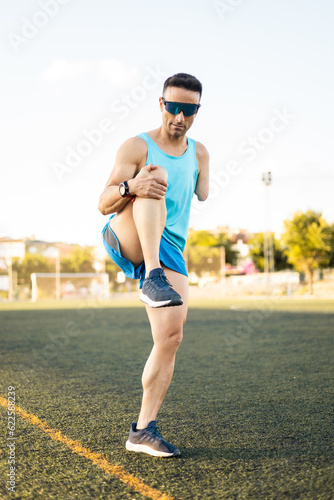 A soccer player between 40 and 50 years old with an amputated arm is stretching his leg standing in the middle of the soccer field. The man is wearing glasses and sportswear.