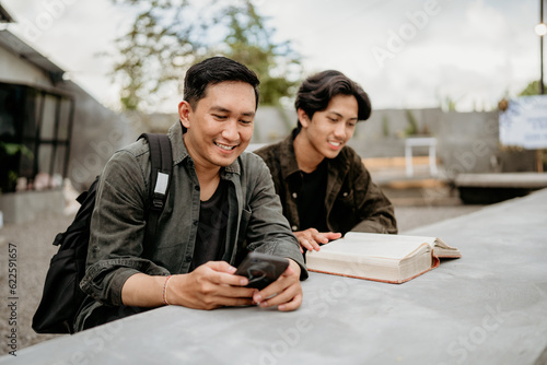 two Asian college students sit using cell phones and read books at an outdoor cafe