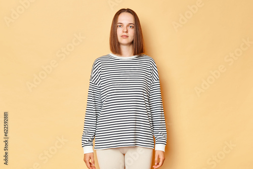 Serious strict brown haired woman wearing striped shirt standing isolated over beige background looking nat camera with bossy stressed expression, being in bad mood. photo