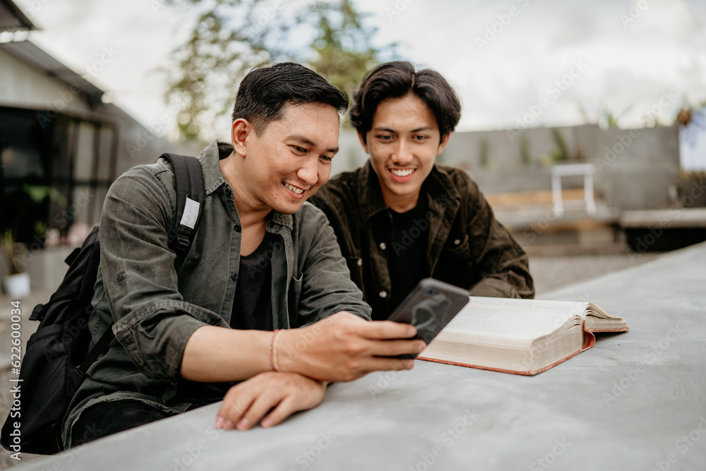 two students sit using their cell phones and read a book at an outdoor cafe two students sit using their cell phones and read a book at an outdoor cafe
