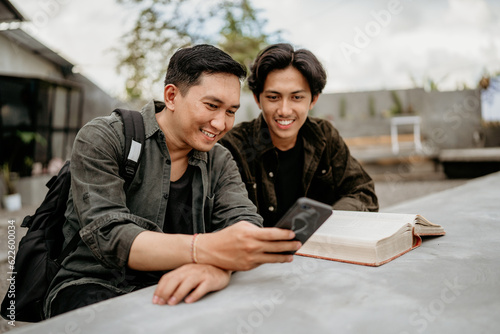 two students sit using their cell phones and read a book at an outdoor cafe