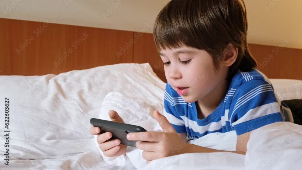 Portrait of a smiling boy playing online video game on a smartphone, lying in bed at home. Happy winner emotions. The kid wins and jumps. Child is using a mobile phone. Concept of gambling addiction. Portrait of a smiling boy playing online video game on a smartphone, lying in bed at home. Happy winner emotions. The kid wins and jumps. Child is using a mobile phone. Concept of gambling addiction.