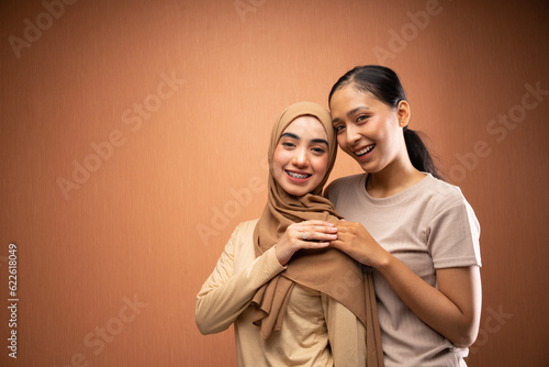 happy women hijab and no hijab wearing cream t-shirt hugging each other and smiling in orange background © Odua Images