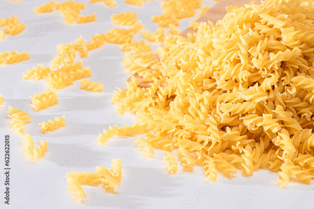 Fototapeta premium Uncooked Fusilli Pasta Scattered on Wooden Board and White Table. Raw and Dry Macaroni. Fat and Unhealthy Food