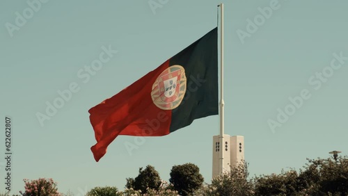 A medium telephoto view of the giant flag of Portugal waving in the wind on a cloudless warm sunny day above a line of green trees and bushes; a thick metal flagpole; slow motion