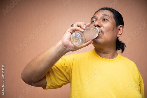 old asian man holding and drinking from unlabelled plastic bottle in plain background isolated
