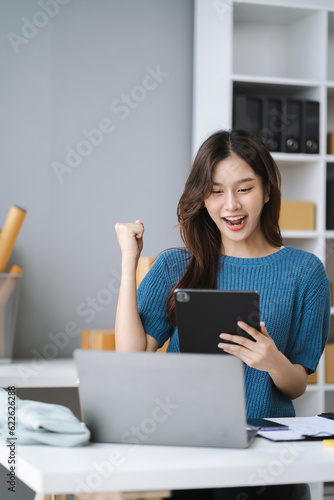 Happy young Asian woman entrepreneur, Smile for sales success after checking order from online shopping store in a smartphone at home, Concept of merchant business online and eCommerce.