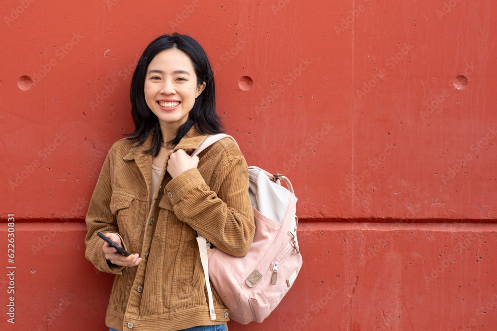 smiling young woman standing with mobile phone and bag by wall smiling young woman standing with mobile phone and bag by wall
