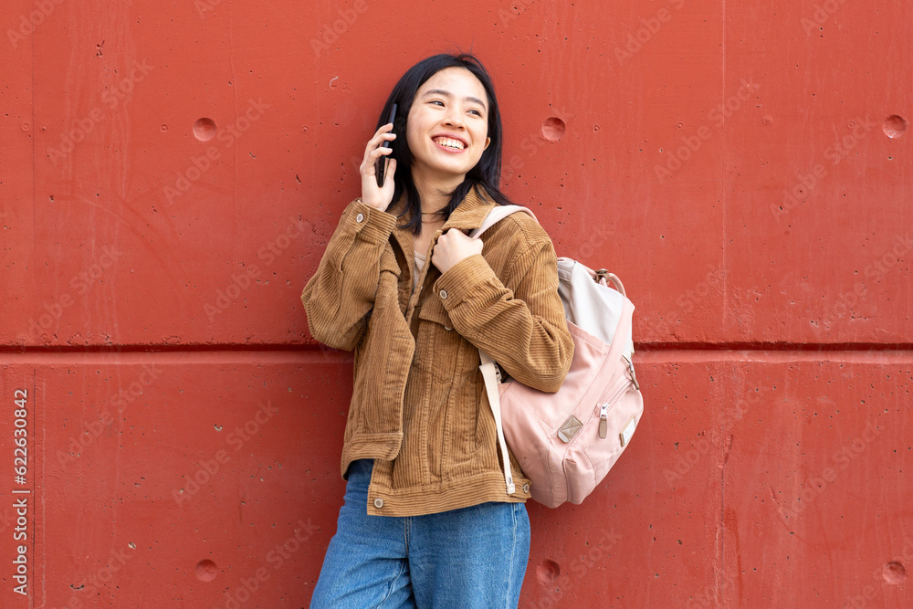 smiling young woman standing with mobile phone and bag by wall smiling young woman standing with mobile phone and bag by wall