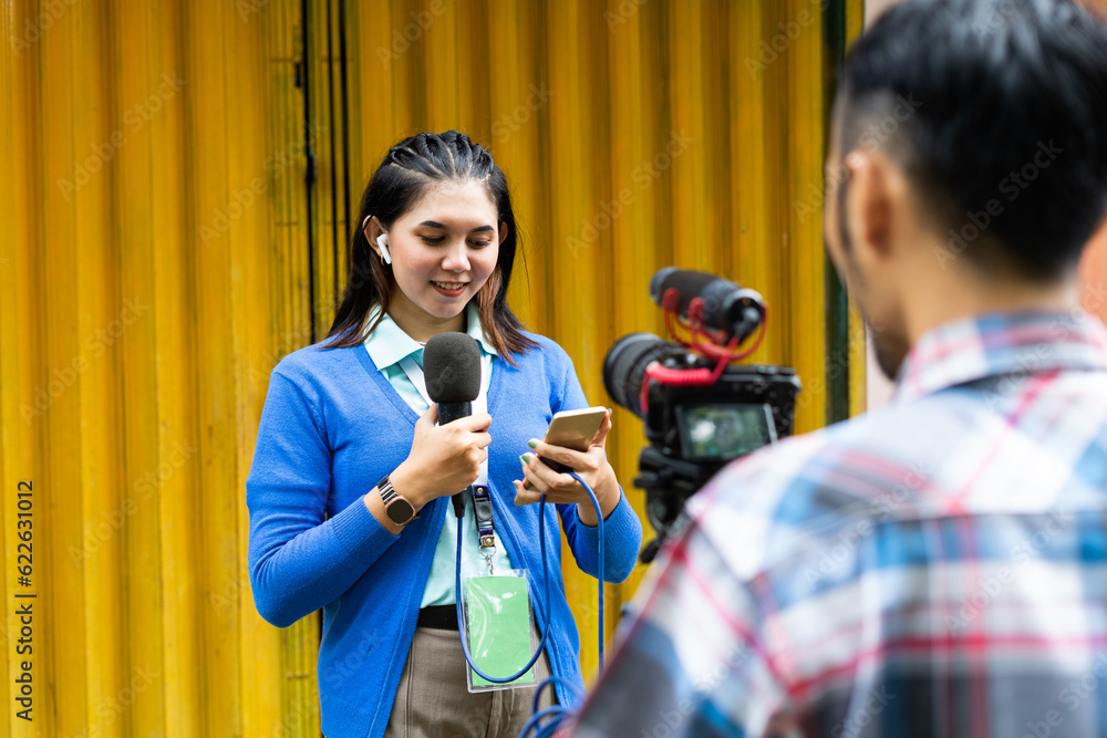 Naklejka premium a beautiful reporter in blue cardigan look at her phone while recording videos for news material with the cameraman using one set of camera and a black microphone