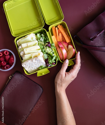 Foto Female hand holding lunch box with sandwiches, carrot sticks, apple, lettuce and raspberries on a burgundy background with a woman's handbag and wallet
