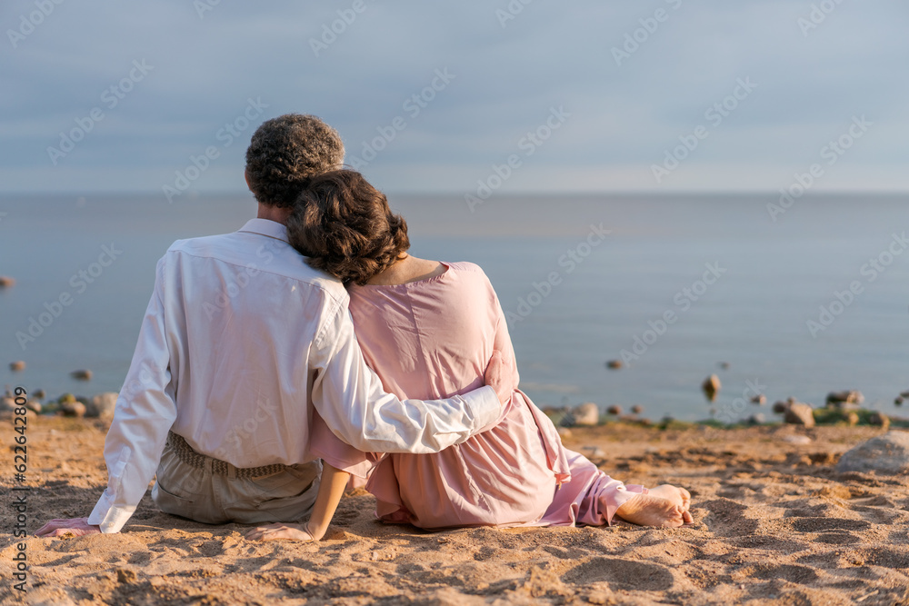young woman in pink dress and an elderly man sit hugging together on beach at sunset. concept of the relationship of different ages in a couple