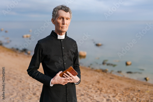 a mature man in a black cassock on the seashore at sunset with a bible in his hands