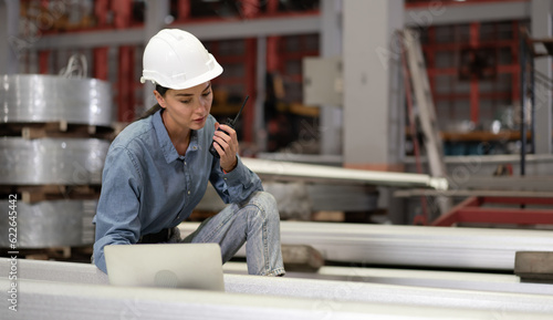 Factory female worker using laptop computer checking, talking to supervisor in heavy metal production facility. Woman technician in white hardhat working, inspecting quality control in manufacturing.