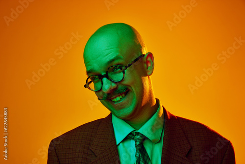 Portrait of young bald man with stylish moustache wearing glsses, tie and suit, smiling against orange studio background in neon light. Concept of human emotions, facial expression, lifestyle photo