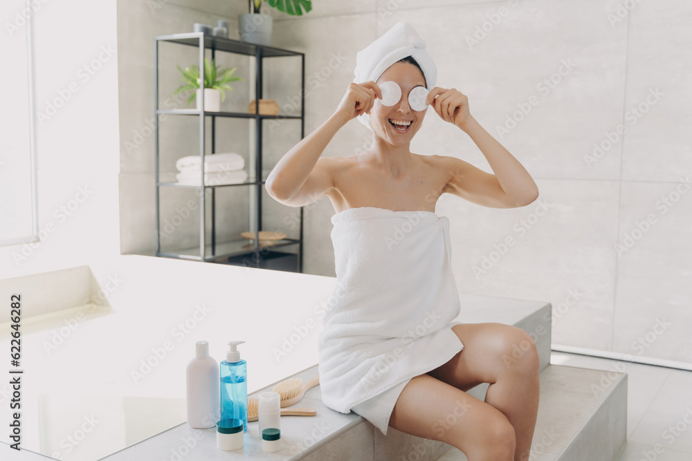 Happy woman applies face toner, has fun with cotton discs. Gorgeous European girl, towel-wrapped post-bath. Lady relaxes on bathtub, showers at home. Happy woman applies face toner, has fun with cotton discs. Gorgeous European girl, towel-wrapped post-bath. Lady relaxes on bathtub, showers at home.