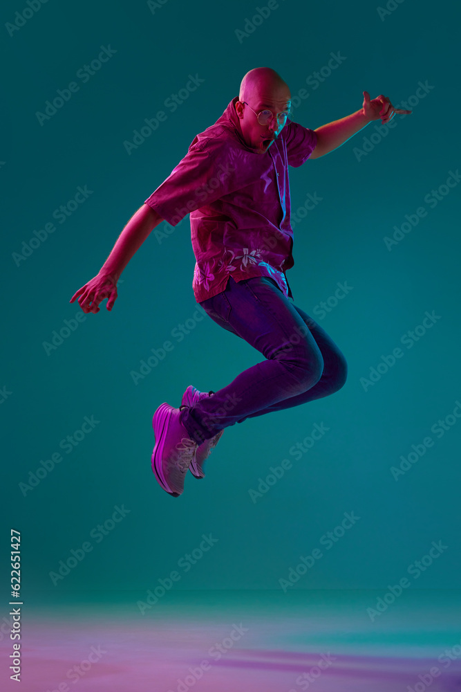 Full-length portrait of young bald man with moustache in stylish clothes emotionally jumping against cyan studio background in neon light. Concept of human emotions, facial expression, lifestyle