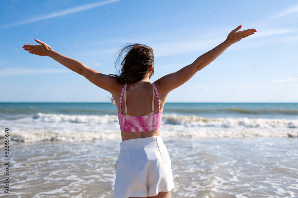 Beautiful adult brunette woman in a pink top and white shorts on a sandy seashore rejoices in travel and vacation Beautiful adult brunette woman in a pink top and white shorts on a sandy seashore rejoices in travel and vacation