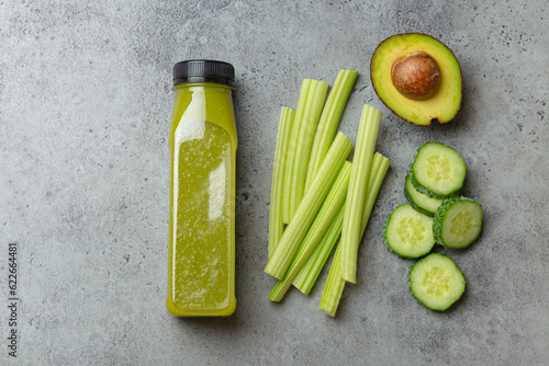 Bottle of green vegetable smoothie with fresh celery sticks, cut avocado, cucumber on gray concrete background top view. Diet beverage, healthy nutrition, detox cocktail