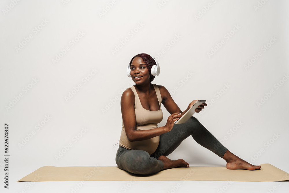 Fototapeta premium African pregnant young woman in headphones with tablet sits on mat