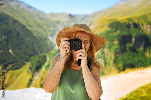 Portrait of a smiling happy elderly woman tourist traveling with a camera posing against the backdrop of mountains. Old elegant lady in straw hat on grass at countryside. Active retired people concept
