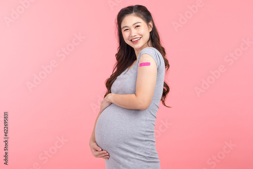 Pregnant Asian Woman smile and happiness after received anti virus vaccine cheerful with bandage isolated on pink background,Pregnancy of young woman enjoying with safety life after got vaccination